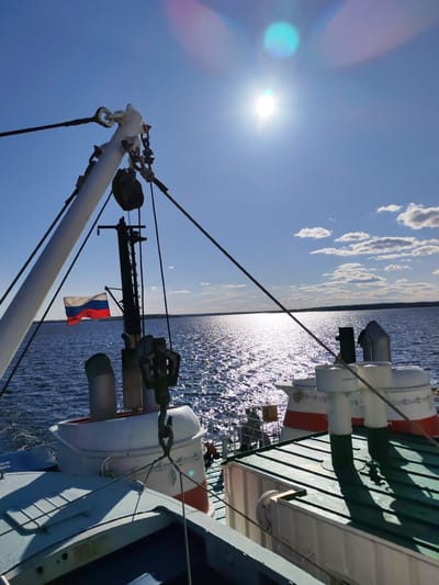 The image shows a view from a ship on a sunny day, with a clear blue sky and sunlight reflecting on the water. The ship features a white boom with rigging, a mast with a Russian flag, and various deck structures, including vents and hatches. The city of Береговой, Russia, is not visible in this image. The environment consists of open water and distant land.