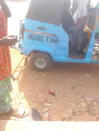 The image captures an outdoor scene in Lapai, Nigeria, featuring a light blue motorized tricycle, commonly known as a "keke napep," positioned on an unpaved, reddish-brown dirt surface. The tricycle has a black soft top and displays the markings "DELUXE" above "NGSIRS 7760" in black stencil-like font on its side. Its rear wheel, equipped with a green hubcap, is visible. Inside the tricycle, partially visible, are at least two individuals; one is dressed in dark clothing, and another wears a white head covering and a patterned garment. A black bag is also visible within the vehicle. To the left of the frame, a person is partially visible, wearing a red/orange patterned top and a green/blue patterned skirt, with a light-colored sandal on their right foot. Their hands appear to be manipulating a small, dark object. The ground is littered with various debris, including a crushed red beverage can, silver-colored wrappers, and small wood fragments. The scene is illuminated by strong, direct sunlight, casting shadows.