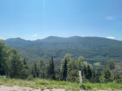 The image depicts a wide, elevated view of a lush, forested mountain range under a clear, bright blue sky. In the foreground, a grassy embankment slopes down towards a gravelly surface, suggesting a roadside perspective. A white and grey striped pole, likely a road marker, stands vertically on the right side of the frame, partially obscured by foliage. The middle ground is dominated by dense, verdant forests covering the rolling hills and mountains. Patches of lighter green, indicating fields or clearings, are visible nestled within the trees in the distance. A few wispy clouds are scattered across the expansive sky. The overall impression is one of a serene, natural landscape on a sunny day in the vicinity of Podkraj, Slovenia, a region known for its picturesque mountainous terrain.