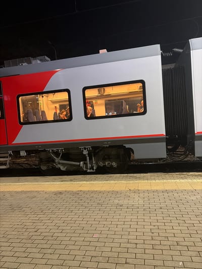 The image shows a side view of a train parked at a station platform. The train cars are white and red, with several windows visible. Inside the windows, people can be seen sitting. The platform is paved with bricks, and a yellow tactile paving strip is visible along the edge. The scene is illuminated by artificial light, suggesting it is nighttime or early morning. The train appears to be a passenger train, and the environment suggests a public transportation setting in the city of Kirpichnoye, Russia.