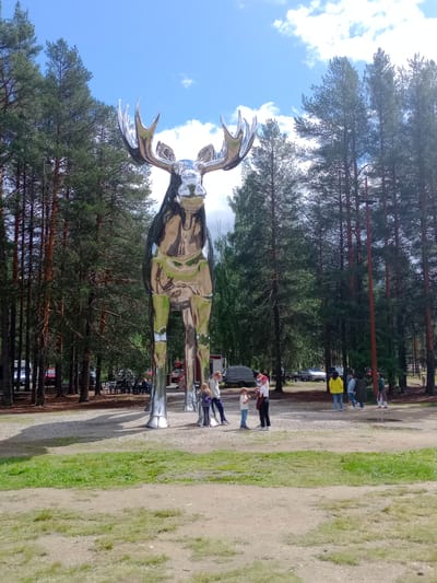 In Atna, Norway, a massive, highly polished chrome sculpture of a moose stands tall amidst a backdrop of tall pine trees and a partly cloudy blue sky. The sculpture, reflecting its surroundings, occupies the central focus of the image. At the base of the sculpture, a group of people are gathered, appearing to admire it. Among them are children and adults, some interacting with each other and others looking towards the sculpture. The ground around the sculpture is a mix of gravel and sparse green grass. In the background, cars and picnic tables are visible, suggesting a public park or gathering area. The overall scene depicts a unique art installation in a natural setting.
