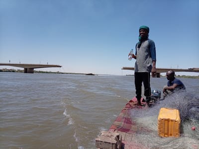 Two men are on a small boat traveling on the Nile River in Bahri, Sudan. A dark-skinned man wearing a green head covering, grey long-sleeved shirt, and dark pants is standing on the bow of the boat, holding a plastic water bottle. Another dark-skinned man wearing a blue shirt is sitting on the boat next to him. A pile of fishing nets and a yellow rectangular container are on the boat. A small outboard motor is attached to the boat. The river is wide and brown with ripples. In the background, there is a damaged bridge spanning the river. The sky is clear and blue.
