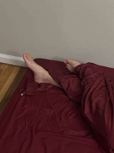 The image captures an indoor scene, likely a bedroom, from an elevated perspective looking downwards. The primary subjects are the bare feet and lower legs of a person, partially covered by dark red or maroon bedding. One foot, with its toes and part of the top visible, emerges from the covers near the upper right of the frame. The other foot and a portion of the lower leg, displaying light skin and some sparse hair, extend further out from under the bedding towards the bottom left, resting close to a light gray wall.
The bedding itself is a solid dark red or maroon color, exhibiting a subtle vertical striped pattern and appearing rumpled, suggesting a person is either resting within it or has recently been in the bed. This bedding occupies the right side of the image. The left side is dominated by a plain, light gray or beige wall, which shows minor imperfections on its surface. At the bottom of the image, a section of wooden floor is visible, along with a white baseboard marking the transition between the wall and the floor.
The activity depicted is a person resting or sleeping, with their feet exposed from the covers, a common position for comfort. The scene suggests a private, relaxed, and domestic setting. There are no direct indications of the time of day, weather, or explicit emotions. No text is visible within the image. The context places this scene in Franklin Township, United States of America.