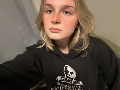 A young woman with blonde hair and fair skin wears a black sweatshirt with a graphic print. The graphic features the word "SKATES" above a skull-like image. Her hair is styled with loose waves and a side part. She has a nose piercing and a small earring. The background is a light, neutral tone with a hint of what appears to be a green pillow or blanket in the upper left corner. The image is vertically oriented, with the woman's face centered.