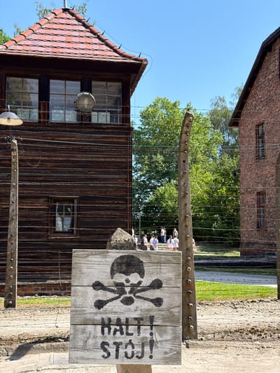 The image shows a wooden sign with a skull and crossbones symbol and the text "HALT! STEH!" (German for "Halt! Stand!") painted on it. The sign is weathered and appears to be made of several planks of wood nailed together. It is mounted on a wooden post, and in the background, there are barbed wire fences and brick buildings. The scene suggests a historical site, possibly a former prison or concentration camp. The sky is clear and blue, indicating a sunny day. The overall impression is somber and historically significant.