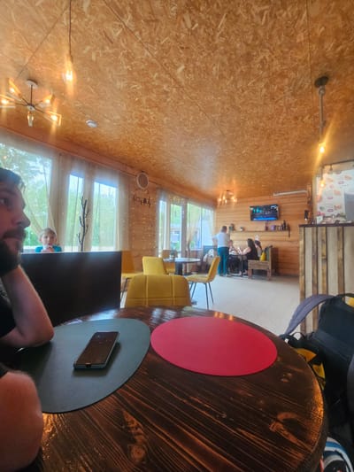The image shows the interior of a cafe in Речицы, Russia. The cafe has a wood-paneled ceiling and walls, and large windows that look out onto greenery. There are several people in the cafe, including a child visible through the window, and other patrons seated at tables in the background. A round, wooden table dominates the foreground, with red and grey placemats and a smartphone placed on the latter. A man's face and shoulder are visible in the lower center, suggesting he is seated at the table and took the photo. In the background, a television is mounted on the wall, and there are yellow chairs and tables, indicating a casual dining area.
