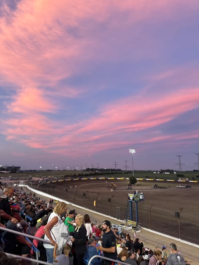 An eye-level, medium shot captures a dirt racetrack at dusk, with a vibrant pink and purple sky overhead. Spectators fill the bleachers in the foreground, some facing the track and others conversing. The track itself is empty except for a few utility vehicles, a starting flag, and a victory lane platform. In the distance, stadium lights, power lines, and distant buildings suggest an urban or suburban setting. The overall impression is one of a paused moment at a sporting event, bathed in the dramatic light of sunset.