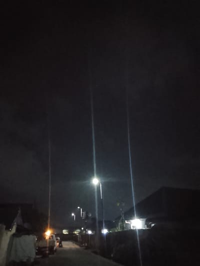 A street scene at night in Rumuokwuta, Nigeria, is depicted in this image. The sky is dark, with faint clouds visible. Several streetlights illuminate the scene, casting long light beams. In the foreground, a white truck is parked on the left side of the road, with its headlights on and an orange light visible on its roof. A tarp-covered structure is next to the truck. Further down the street, more houses and lights are visible. The road appears to be paved.
