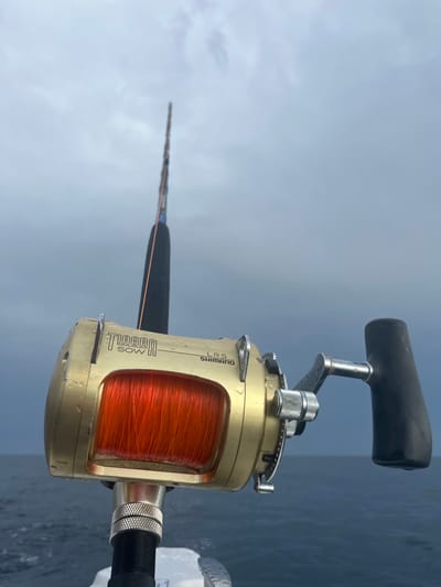 A close-up captures a gold Shimano Tiagra 50W fishing reel filled with orange line, mounted on a black fishing rod. The reel's handle is black. The background is a blurry mix of the sky with overcast, and the ocean. The reel's details, like the lettering and the line winding around the spool, are clearly visible.