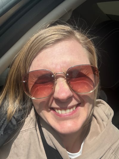 A Caucasian woman with blonde hair and fair skin is smiling while wearing rose-tinted sunglasses. She is wearing a beige hooded sweatshirt and possibly seated in a car. The exact location within Mar'evka, Russia, cannot be determined from the provided image.
