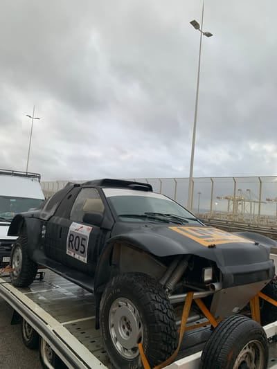 A black, heavily modified off-road rally vehicle, bearing the racing number "R05" on its driver's side and partially visible yellow "MACHINED.B.Y.T.L.E." lettering on its hood, is secured with orange ratchet straps to a white flatbed tow truck. The rally vehicle features prominent exposed front suspension components, large knobby BFGoodrich All-Terrain T/A tires on silver multi-spoke rims, a roof scoop, and a white sun visor strip above the windshield. A white van with a roof rack is partially visible to the left, behind the rally car. The scene is located in Ksar El Majaz, Morocco, in a port or industrial area, evidenced by multiple large gantry cranes visible over a body of water in the background, separated by a fence. Two tall streetlights are also present. The sky is overcast with heavy cloud cover. No individuals are visible.