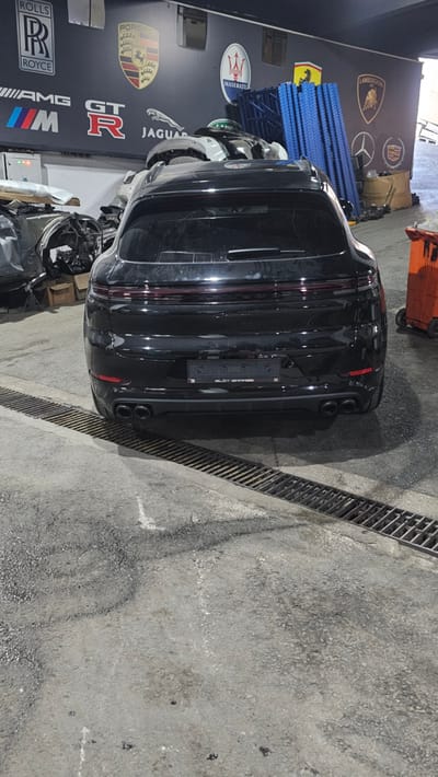 In Ataşehir, Turkey, a black Porsche Panamera is parked in what appears to be an auto repair shop or a storage facility. The car is viewed from the rear, facing away from the camera. The vehicle is positioned over a drainage grate that runs across the concrete floor. In the background, the wall is decorated with various automotive brand logos, including Rolls-Royce, Porsche, Jaguar, Maserati, Ferrari, Lamborghini, Audi, and Cadillac. Parts of other vehicles and disassembled car components are visible on the left side of the image, along with stacked blue metal shelving units and an orange dumpster on the right. The lighting suggests an indoor, possibly industrial, environment.