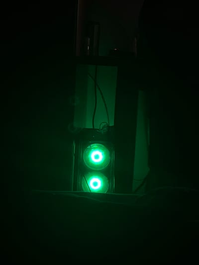 The image depicts a dimly lit room, possibly a bedroom or office, with a focus on a computer tower that features two illuminated green fans. The computer tower is placed on a surface, possibly a desk or shelf, in the center of the frame. The green glow from the fans casts an eerie light on the surrounding dark environment. To the right of the computer, there is a tall, dark object that could be another piece of furniture or a wall. A black wire snakes from the top of the computer tower, disappearing into the darkness. The overall impression is one of a technologically focused space, possibly used for gaming or computing, with a distinct aesthetic created by the green lighting. There is no indication of any specific location in Skawina, Poland, within the image itself.