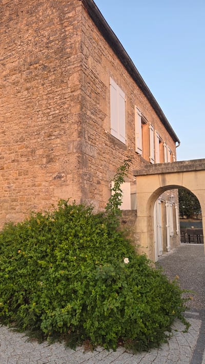 A stone building with white shutters stands at an angle, partially obscured by a lush green bush with small yellow and white flowers. The building's facade is constructed from rough-hewn, tan-colored stones. Sunlight casts a warm glow on the stonework. An arched entryway with a cobblestone path leads into the building's interior, where more white shutters are visible. The sky above is a clear, pale blue. The setting appears to be a serene and historic location in the city of Macé, France.