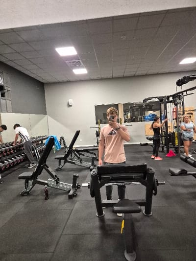 The scene is an interior view of a fitness center or gym, located in San Lorenzo de El Escorial, Spain.

**People:**
*   **Central Figure (Reflected):** A male with light brown, curly hair stands centrally, holding a dark smartphone in his right hand, capturing his reflection in a large mirror. He wears a light peach-colored short-sleeved t-shirt with a dark graphic on the chest and dark trousers. His lower legs are obscured by equipment.
*   **Left Background:** Two males are present near a dumbbell rack. One, wearing a white t-shirt with dark horizontal stripes and dark pants, is reaching for a dumbbell. The other, mostly obscured, wears a white t-shirt.
*   **Right Mid-ground:** A female with dark hair pulled back, wearing a black tank top, black leggings, and bright pink athletic shoes, stands near a multi-station cable machine with her left arm slightly raised.
*   **Right Background:** Another female, with lighter hair, wearing a light blue short-sleeved top and light-colored shorts, stands further right near the same multi-station machine.

**Objects and Equipment:**
*   **Foreground:** A black exercise machine, consistent with a preacher curl or similar arm isolation unit, featuring a padded seat and armrest, is directly in front of the viewer.
*   **Left Mid-ground:** Two black adjustable utility benches are present. The front-most bench has a blue towel draped over its backrest, and a dark-colored bottle rests on the floor underneath it.
*   **Left Background:** A multi-tiered rack filled with numerous black hexagonal dumbbells of various weights.
*   **Right Background:** A black multi-station cable machine or squat rack with integrated weight stacks and various attachments. Red conical markers are visible on the floor near this equipment.
*   **General:** The floor is covered with black interlocking rubber mats. The walls are painted a uniform light gray. The ceiling is a suspended acoustical tile system with several square-shaped recessed fluorescent light fixtures providing bright, even illumination. A large mirror occupies a significant portion of the background wall.

**Actions and Interactions:** The central male subject is actively taking a mirror selfie. The other individuals appear to be engaged in independent exercise routines or are resting within the gym environment. No direct interaction between individuals is discernable.