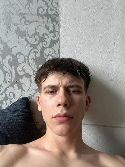 A young man is photographed from the chest up, looking directly at the camera. His head is slightly tilted, and his expression is neutral. He has short, dark, wet hair and appears to be in a bedroom, with a patterned wallpaper on the left side of the frame and a textured white wall on the right. A dark blue pillow is visible to his left. The lighting suggests it is daytime, with natural light illuminating his face. No specific location in the city of Hamm, Germany can be determined from the visual information provided.