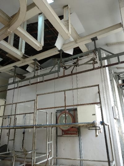 The image depicts the interior of an industrial or commercial facility located in the city of Banjar Wangi, Indonesia.

The environment features a white panel ceiling with multiple sections dislodged, damaged, or missing, revealing a dark, open cavity above containing exposed structural beams and conduits. White-painted steel beams, part of the building's structure, are prominently visible in the upper portion, with some appearing to support or connect to an overhead gantry system. The walls are constructed from light-colored, possibly insulated, panels.

A complex network of industrial equipment dominates the scene. In the upper half, an overhead gantry or conveyor system, composed of dark and light-colored metal rails and support structures, traverses the space. Below this, a multi-tiered metal framework or scaffolding, constructed from square and round metal tubing, occupies the mid-ground and foreground. This structure exhibits signs of rust and wear, featuring numerous horizontal bars and vertical supports. A blue rope is draped over one of the horizontal bars in the mid-ground, and a white cloth or towel hangs from a bar on the right side of this structure. A metal hook is suspended from a lower section of the metal framework on the left.

Mounted on the far wall, at approximately mid-height, is a large, rectangular industrial scale with a prominent circular red housing and a green dial face. To the immediate right of the scale, a white rectangular electrical box or control panel is affixed to the wall.

No individuals are present in the image, and no active actions or interactions are depicted; the scene is static.