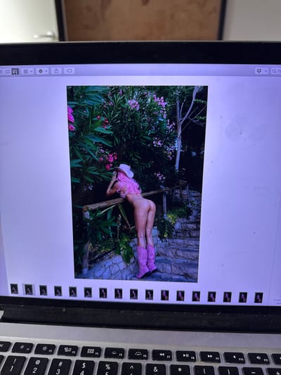 The media file displays a digital image on a laptop screen, featuring a person posing in an outdoor setting. The subject is a person with pink hair, wearing a pink cowboy hat and pink boots, with a light-colored bikini bottom. They are lying on their side, propped up by their elbow, amidst lush green foliage and pink flowers. A rustic wooden structure, possibly a ladder, is visible behind them, leaning against a stone wall. The overall scene suggests a natural, possibly garden or park-like environment. The image is displayed in a photo editing or viewing application on a MacBook laptop, indicated by the application interface elements visible on the left and the keyboard on the right. The time of day appears to be daytime, with natural lighting illuminating the scene. The crops provided offer closer views of the subject, the surrounding vegetation, and the laptop interface, including small thumbnail previews of other images along the right side of the screen. The visible text on the laptop screen is limited to interface elements, including icons, and some Russian text in the header area which translates to "Show" and "In". The location context provided is Mandelieu-la-Napoule, France.