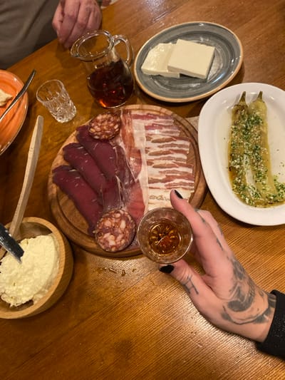 A table setting displays a charcuterie board with various cured meats, sausage, and bacon. Adjacent are plates of cheese and sauteed peppers. A person holds a shot glass of amber liquid, and a small pitcher of a similar liquid is also visible. A bowl of whipped cream or soft cheese sits next to a wooden spoon and knife. The table is made of wood.