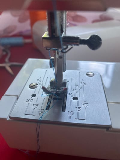 This is a close-up, overhead shot of a sewing machine needle and presser foot with blue thread. The needle is lowered and threaded through the eye of the presser foot, with excess thread trailing down. The presser foot is positioned over the metal stitch plate of the sewing machine, which has inch and centimeter markings. The background is blurred and shows glimpses of other sewing supplies, including spools of thread and scissors. The scene appears to be indoors, likely in a home or workshop setting. There are no people or animals visible in the image, and no specific location in Orlivshchyna, Ukraine can be identified from the visual information provided.