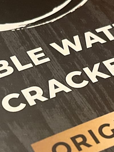 This close-up image captures a section of product packaging, likely a label, featuring prominent text against a dark, textured background. The primary background is a deep charcoal or black with subtle horizontal brushstroke or distressed patterns. On the left, a partial, thick white outline of an irregular circular or oval shape is visible, suggesting a logo or design element.
The text is large, bold, and primarily in white, set at an angle, rotated approximately 30-45 degrees counter-clockwise. Clearly visible words include "BLE WATER" on the left and "CRACKER" in the center. Further to the right and slightly higher, a section of the label is light brown or tan, containing darker, bold text that reads "ORIG". Combining these fragments, the full text likely refers to "TABLE WATER CRACKER ORIGINAL".
No people or activities are present; the image focuses solely on the textual and design elements of the packaging. The lighting is even, suggesting an indoor setting. The clear resolution allows for easy readability of the visible text. The context indicates the item is located in Montgomery, United States.