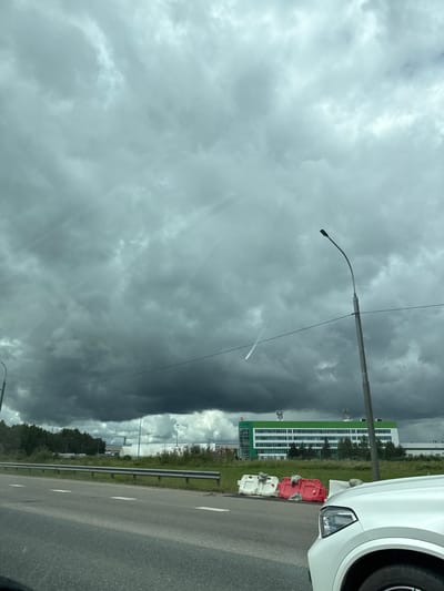 This media file depicts a street scene in Davydovskoe, Russia, with a focus on the sky and its dramatic weather patterns. The primary subjects include a street light pole, a white car on the right side of the frame, a modern building with green and white accents in the background, and a road. The weather is a prominent element, with dark, ominous storm clouds dominating the sky. The clouds are heavy and suggest an impending storm or the aftermath of one, creating a sense of drama and perhaps unease.

The activity is not explicitly depicted, but the image appears to be taken from a moving vehicle, possibly on the road. The presence of traffic barriers (red and white plastic barriers) on the side of the road suggests construction or some form of temporary road management.

Notable details include the time of day, which seems to be daytime, but the heavy cloud cover creates a subdued light. The location cues are the building design and the Russian context provided. The overall emotion evoked by the image is one of atmospheric intensity and the power of nature, with the stormy sky contrasting with the man-made structures. There is no visible text in the image.