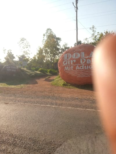 An orange rock painted with "DOLA Gir Dadi Mit Aduga" sits beside a dirt road. A small, gray rock nearby is marked with "HON ODIENO GWN 2021." A road runs horizontally at the bottom. Green trees rise in the background, partially obscured by sunlight. The top of a utility pole is visible. A large, blurry pink object is in the foreground on the right.