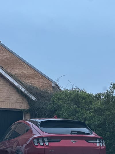 The image displays the rear of a red Ford Mustang Mach-E, featuring its distinctive taillights, the Mustang emblem, and "AWD" badging. The vehicle is parked in Gillingham, United Kingdom, directly in front of a residential garage with a dark door. Behind the garage, a house constructed of light brown brick is visible, with a dark tiled roof. Extensive dark green and bare thorny vegetation covers parts of the garage structure and extends onto the lower section of the house's roofline. Additional dense green foliage occupies the middle ground to the right. The sky overhead is a uniform, light blue. No individuals are present in the scene.