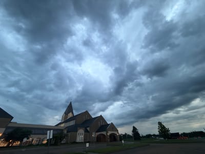 An exterior, low-angle, wide shot depicts the dark, overcast sky above a church building in Stony Plain, Canada. The sky is filled with dense, grey clouds that partially obscure the light. The church building, constructed of brick, features a tall, pointed steeple topped with a cross. A portion of a larger, modern wing with large windows is visible to the left of the main church structure. The foreground consists of a paved area and a grassy verge. There are no visible people in the image. The overall atmosphere is somber due to the weather conditions.