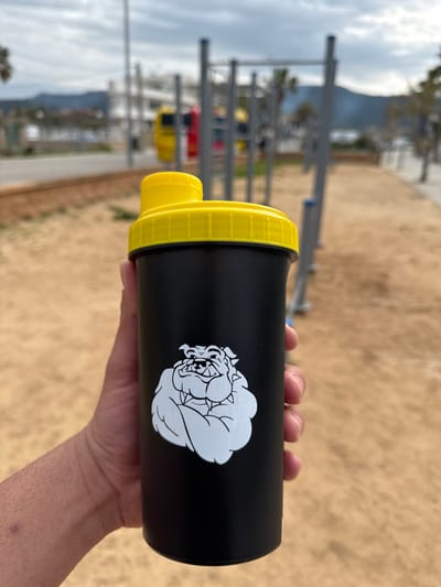 A right hand holds a matte black shaker bottle with a bright yellow screw-on lid and a yellow flip-top spout, positioned centrally in the foreground. A white graphic depicting a muscular bulldog with a spiked collar is affixed to the side of the bottle. The background is an out-of-focus outdoor scene in Port D'andratx, Spain. It features a sandy or dirt ground in the mid-ground with metal calisthenics equipment consisting of various bars and frames. Beyond the fitness equipment, a paved path is visible, followed by a blurred yellow and red vehicle. A body of water, consistent with a harbor, is discernible to the right, with distant mountains under an overcast sky. Scattered palm trees and other sparse vegetation are present in the background.