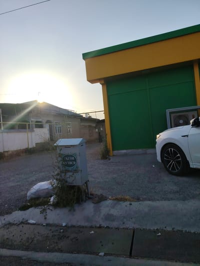 This image depicts an outdoor scene in Pargos, Uzbekistan, with a focus on a building with a yellow and green facade and a white car parked nearby. The sun is low in the sky, casting a warm glow and creating a lens flare effect. In the foreground, a grey mailbox is visible with decorative green lettering. A white garbage bag lies on the ground next to the mailbox. Beyond the building, a residential structure with a corrugated roof is visible, with evidence of wear and tear. The ground is covered in gravel and dirt. A portion of a white car, including its front wheel, is visible on the right side of the frame, parked on the gravel surface. The sky is clear blue with the sun positioned towards the left, creating a hazy atmospheric effect. There are no people clearly visible in the image.