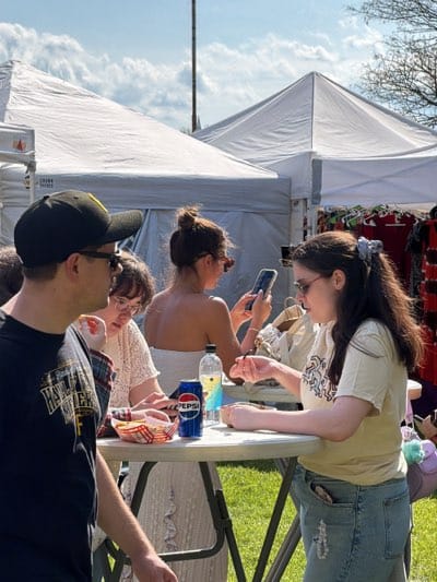 The image shows a group of people gathered around a small round table at an outdoor event, likely a festival or market, in Rochester, United States. The setting appears to be a grassy area with white tents in the background, suggesting a vendor or exhibition space.
The group consists of at least five people:
1. A man in a black baseball cap and t-shirt with sunglasses facing left.
2. A woman with brown hair and glasses wearing a white top.
3. A woman with her hair in a bun, wearing sunglasses and a white top, holding a smartphone.
4. A woman with long dark hair and glasses wearing a light yellow t-shirt.
5. A person standing further to the left of the first man.
The people are standing around a round, white folding table, and there is a basket of french fries, a can of Pepsi, and a water bottle on top. At least one of the woman is eating with a fork, and another is looking at her phone. The woman in the bun is holding a cell phone.
The weather looks bright and sunny, with blue sky visible.
