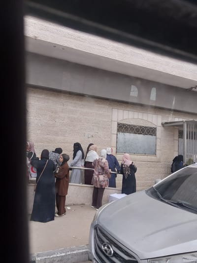 A line of seven women, some wearing headscarves or hijabs, are standing on a sidewalk outside a building in al-Qarara, Palestinian Territories. The building has a light-colored stone exterior with a small, barred window. A silver SUV is partially visible to the right of the women. The viewpoint is looking down at the scene.