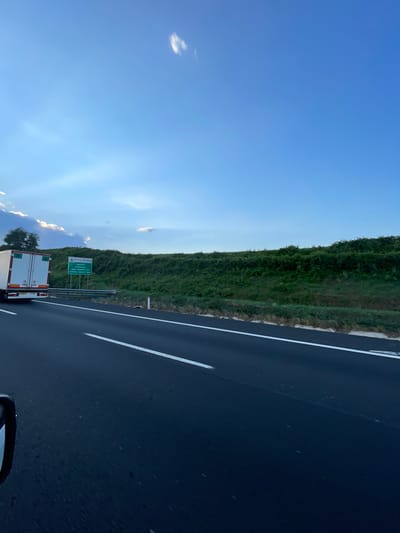 A daytime image depicts a multi-lane asphalt highway under a clear blue sky with scattered white clouds. The perspective is from a vehicle moving on the highway, with a portion of its left side mirror visible in the bottom-left corner. Ahead in the left lane, a white semi-trailer truck is visible from its rear, displaying red markings and green text on its back doors. To the left of the highway and behind the truck, a green road sign with white text is present, indicating "4 Ferma Strada" with a left-pointing arrow, and further text identifying "Paliano," "Arce," and "zona industriale." The right side of the highway features a metal guardrail separating the road from a densely vegetated green embankment that rises upwards. The overall scene suggests a highway in the vicinity of Paliano, Italy, specifically approaching an industrial zone exit referenced on the sign.