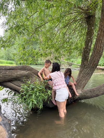 Three individuals, two children and one adult, are positioned on a large tree trunk that extends over water. The water appears to be a shallow river or stream. The scene is heavily vegetated with a variety of greenery, including trees and smaller plants. The tree's branches droop. The children are fair-skinned, one wearing a blue and pink garment, and the other topless. The adult is wearing a pink top and white shorts. The photograph suggests a natural environment, typical of riverside or lakeside scenery. Given the request for location, and based on the description of the scene, the location is the city of West Oxfordshire, United Kingdom.