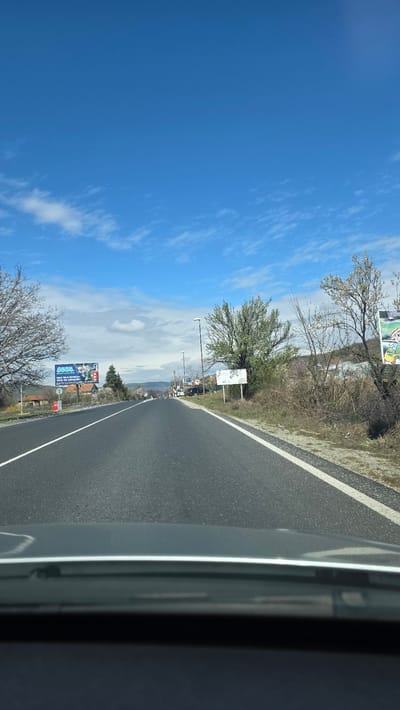 Observed from inside a vehicle traveling on a paved road, the view encompasses a clear blue sky with scattered clouds above and to the left. Adjacent to the road is a grassy verge dotted with trees and vegetation. Signage is visible, including billboards displaying commercial advertising. The road surface is smooth and dark, marked with a white line delineating the edge of the roadway. The scene is in Usoika, Bulgaria.