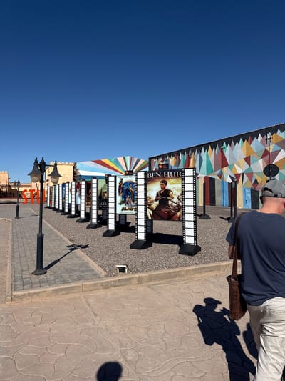 This image depicts an outdoor area within the Atlas Film Studios in Ouarzazate, Morocco, under a clear blue sky. A paved pedestrian pathway, featuring light-colored, patterned tiles, occupies the foreground and left side. A black, ornate streetlight with multiple lamps stands on the left. Behind it, large orange letters spelling "STU" are partially visible. Adjacent to the paved area, a strip of grey gravel extends towards a series of buildings.
Lining the gravel strip is an array of approximately ten vertical display stands, designed to resemble film strips with sprocket holes along their edges. Each stand features a poster for a motion picture. Visible titles include "BEN HUR," "ALADDIN," and "ALEX THE GREAT," with an "ATLAS FILMS" logo also discernible on multiple posters.
In the background, several distinct buildings are present. On the far left, a low, tan-colored structure with crenellated parapets is visible. Further right, a building features a large, fan-shaped facade with radiating stripes in various colors (blue, white, yellow, red, orange). To its right, a long building is characterized by a prominent geometric mural covering its wall, composed of numerous triangular and quadrilateral shapes in an assorted palette including brown, green, blue, pink, and yellow. Dark vertical support beams are visible against this wall, along with dark doorways.
In the right foreground, a male individual is depicted from behind, walking towards the right. He wears a dark blue t-shirt, light-colored pants, and a grey baseball cap. A dark brown leather satchel is slung over his right shoulder. Strong sunlight casts distinct shadows of the man and the streetlights onto the ground.