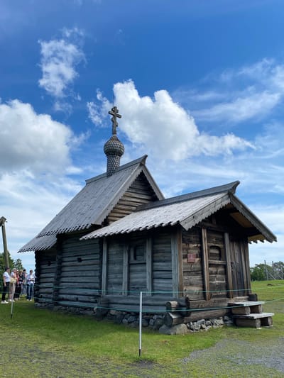 A wooden church stands in the foreground against a bright blue sky with scattered white clouds. The church is constructed from logs and features a multi-tiered roof, with a central dome topped by a cross. The structure has a rustic appearance, with visible wood grain and weathered timber. A small group of people, dressed in casual clothing, are standing to the left of the church, observing it. The ground around the church is covered in green grass, and a low rope barrier is visible in front of the building. The background shows a wooden fence and trees in the distance, suggesting an outdoor, possibly historical, setting. The location is Boyarshchina, Russia.