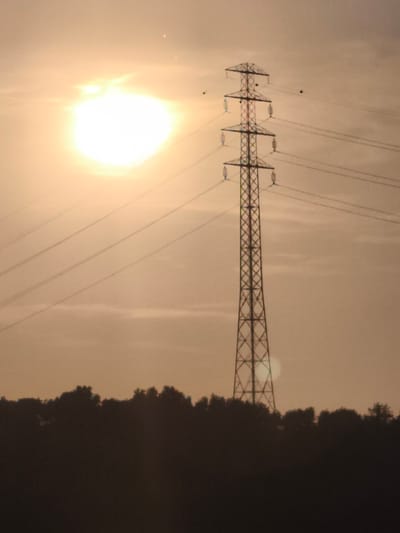 The image captures a silhouette of a high-voltage electricity pylon standing tall against a hazy, sunlit sky. The sun, a bright, diffused orb, is visible in the upper left quadrant of the frame, casting a warm, golden light that permeates the atmosphere. Several power lines stretch diagonally across the frame, originating from the pylon and disappearing into the distance. In the lower portion of the image, a dense line of trees forms a dark silhouette against the lighter sky, obscuring the ground. The pylon itself is a lattice structure, with crossbars and insulators clearly discernible. A few small, dark specks, possibly birds, are visible in the sky near the pylon. The overall impression is one of an industrial landscape at dusk or dawn, with the power infrastructure dominating the scene. The image appears to have been taken from a distance, likely in an outdoor setting.