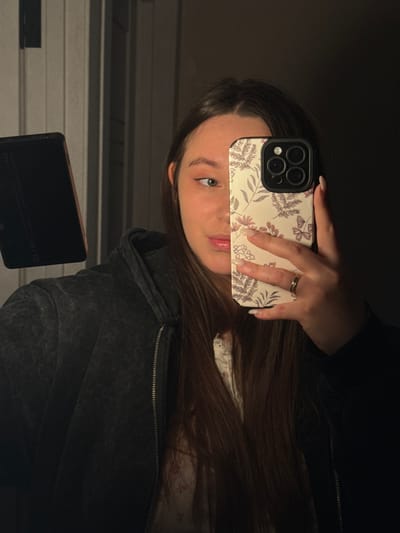 A female subject is depicted taking a mirror selfie in an indoor setting within Prikubanskii, Russia. The individual, possessing long, dark brown hair parted down the middle, is wearing a dark gray zippered hoodie over a light-colored top with a subtle floral pattern. Her left eye, with a slight upward and rightward gaze, is visible, alongside portions of her face showing light pink eyeshadow and lip gloss. Her right hand, adorned with a gold-toned ring on the fourth finger and light-colored manicured fingernails, holds a smartphone vertically, obscuring the right side of her face. The smartphone is an iPhone model featuring a three-lens camera array, encased in a cream-colored protective cover with a repeated pattern of pink and purple floral motifs and butterflies. To the left of the subject, a dark-colored tablet or e-reader, possibly with a cover, is partially visible. The scene is dimly lit, with a localized light source creating reflections on the phone, tablet, and subject's face. The background shows an indistinct, plain wall or doorframe.