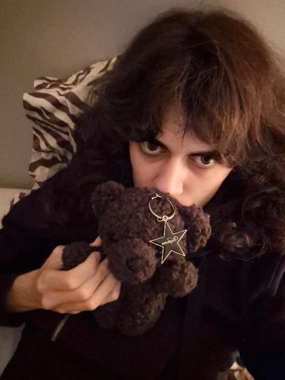 A person with long dark hair and fair skin is holding a brown teddy bear with a star-shaped tag. The person's face is partially obscured by the bear, but their green eyes are visible. The person is wearing a dark-colored top, and a pillow with a brown and white zebra print is visible in the background. The photo is taken from a slightly high angle.