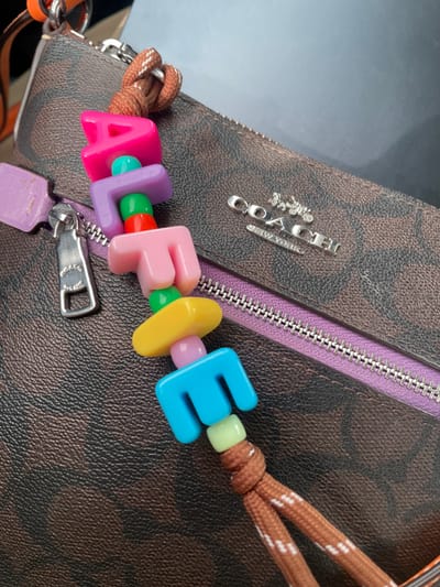 The image presents a close-up view of a brown-patterned bag, likely a purse or tote, made of a textured material with a repeating 'C' monogram, characteristic of the Coach brand. A prominent silver metallic "COACH NEW YORK" logo, featuring a horse and carriage emblem, is affixed to the bag's surface.

A colorful, personalized keychain spelling "ALFIE" is centrally featured, attached to the bag. The letters are distinct: a bright pink 'A', a purple 'L', a light pink 'F', a yellow 'I', and a blue 'E'. These letter beads are separated by smaller, round spacer beads in green, orange, light green, and light purple. The keychain is secured to the bag by a brown braided rope-like strap with white specks.

A purple fabric zipper runs horizontally across the bag, equipped with a silver zipper pull that bears a small, embossed "COACH" logo. Below the zipper pull, a small, light purple rectangular plastic charm is visible. A portion of an orange strap or loop is also seen on the far left, attached to the bag.

The background is uniformly blurred, appearing as a light, possibly reflective, gray surface, offering no specific environmental details. The photograph focuses entirely on the bag and its accessories, presenting them as static objects. There are no people visible, nor is any activity or event depicted. The image solely showcases these branded and personalized items. No visual cues within the image contextually link it to Aratashen, Armenia.