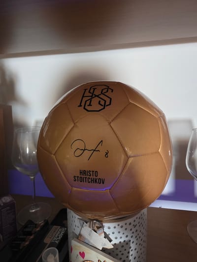 A close-up, eye-level, indoor shot features a golden soccer ball as the main subject, positioned on top of a decorative cylindrical container. The ball displays a black logo with intertwined letters "HS" and a signature that appears to be of Hristo Stoichkov, along with his name printed below in black capital letters. The background includes a white wall and shelf, with two wine glasses on either side of the ball. The bottom portion of the image reveals several boxes and what appear to be banknotes. The lighting is moderate, casting some shadows and highlights on the glossy surface of the soccer ball. The scene is an indoor setting, likely a shelf in a home or office. There is no indication of the city of Rovigo, Italy, in the image.