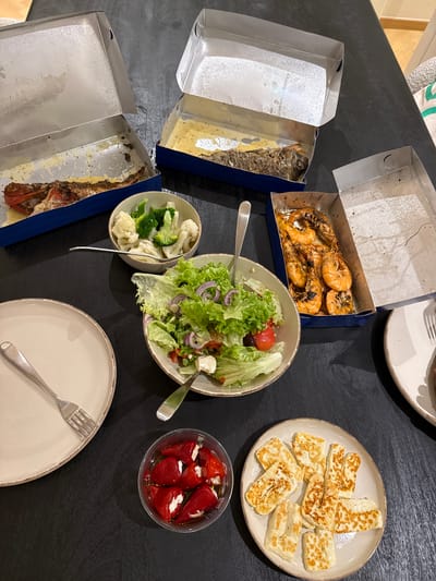 A top-down shot reveals a table laden with a meal, likely in a residence in the Municipal Unit of Elliniko, Greece. Several dishes are arranged: one takeout box containing grilled shrimp, another with fish, a plate of fried halloumi cheese, a salad with greens, red onion, and tomatoes, a small bowl of cooked broccoli and cauliflower, and a clear container of small, red peppers stuffed with a white filling. Plates and forks are also visible, set for dining. The takeout boxes suggest the meal was likely brought in, not prepared on-site.