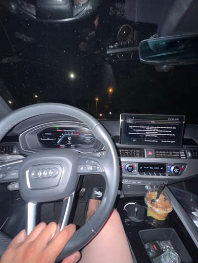 The image shows the interior of a parked Audi car at night. The steering wheel is in the foreground, with the Audi logo visible. The dashboard features a digital display showing a notification about online services and data collection. The car's infotainment system displays climate control settings. A cup with a straw sits in the cup holder. The driver is holding the steering wheel. The location cannot be determined from the image. The location of Schaumburg, United States of America is irrelevant to the context of the image.
