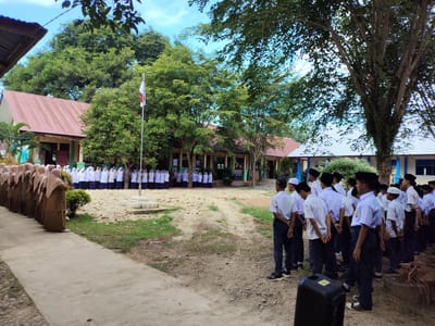 This image depicts an outdoor scene at what appears to be a school in Keudearon, Indonesia. In the foreground, a row of students, predominantly male, stand at attention facing towards the left. They are dressed in uniforms consisting of white shirts, dark pants, and black caps or hats. Some students have dark trousers and white shirts, while others have dark trousers and white shirts with black caps. To the left, a separate group of students, predominantly female, are also lined up, facing the same direction. They are wearing brown headscarves and brown uniforms.

Behind the students, several school buildings with reddish-brown roofs are visible, interspersed with lush green trees. A flagpole with an Indonesian flag (red and white) is positioned in front of one of the buildings. The ground is a mix of dirt, gravel, and sparse grass. In the far right of the image, another group of students in white shirts and dark pants and caps are standing in formation. A black speaker is placed on the ground in the lower right corner of the frame. The sky is visible through the foliage of the trees, with some clouds present. The overall impression is that of a school assembly or flag ceremony.