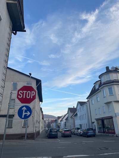 The photo captures a street scene with buildings lining both sides. A stop sign and a directional sign are visible on a pole. Cars are parked along the street. The sky is predominantly blue, filled with wispy clouds. A jet contrail cuts across the upper portion of the sky. The angle of the shot looks upwards, accentuating the height of the buildings.