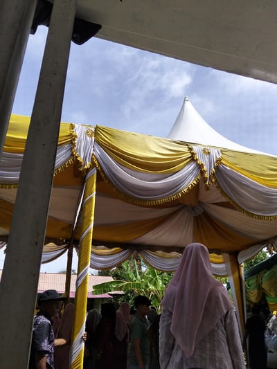 In Cottrueng, Indonesia, a gathering is taking place under a large, decorative tent. The tent's canopy is adorned with flowing yellow and white fabric, accented with gold fringe and a circular gold embellishment at its peak. Supporting the tent are substantial gray concrete pillars, one of which is prominently featured in the foreground.
Several individuals are present. To the left, an elderly man wearing a black patterned shirt and a black cap stands with his side to the camera. He appears to be in conversation or observing the surroundings. Towards the center, a person from behind, wearing a light pink hijab and a patterned garment, is visible. Other individuals in the background are partially obscured by the tent's structure and their attire suggests a formal or cultural event.
Behind the tent, traditional Indonesian housing with red tile roofs and lush green foliage, including banana trees, are visible. The sky above is bright blue with scattered white clouds, indicating daylight. The overall scene suggests an outdoor event, likely a celebration or community gathering, in a semi-rural or residential setting in Cottrueng.