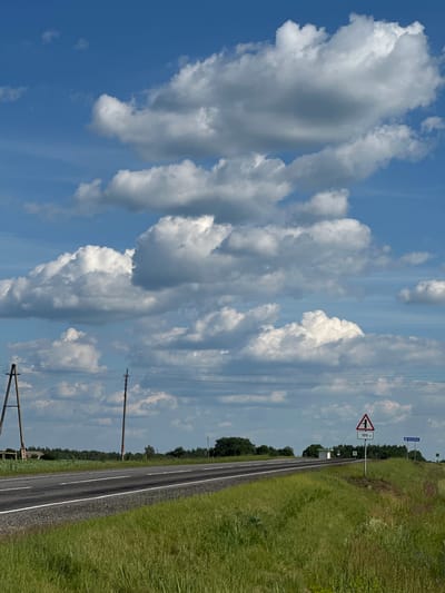 The image depicts a rural roadside scene in Belarus under a bright, partly cloudy sky. The perspective is from a low angle, looking up at the sky and along the road. In the foreground, a strip of lush green grass lines the right side of a paved road with a white stripe down the center. Beyond the road, a dense line of dark green trees forms the horizon.
Several power lines and utility poles traverse the scene, one prominently in the lower right quadrant, and others receding into the distance. A triangular road sign with a red border indicates a sharp turn ahead and reads "100 m" below the symbol. Another smaller, square green sign with a white symbol is visible further down the road. In the distance, a blue road sign with white Cyrillic text reading "СКАЛІНА" is visible, indicating a direction or place name.
The sky is a vibrant blue, filled with large, fluffy white cumulus clouds that cast soft shadows. The overall impression is of a sunny day, likely late morning or early afternoon, with a sense of open space and quiet countryside. There are no people or animals visible in the frame. The scene appears to be a typical stretch of road in a Belarusian rural area, possibly near the village of Skalina.