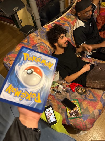 Two young men are lying on a bed with a patterned mattress. One man wears glasses and a black and white shirt, holding a lit joint. The other man has curly hair, a beard, and a black shirt. Beside them on the bed are miscellaneous objects including a red game controller, lighters, rolling papers, an ashtray filled with cigarette butts, a DVD case, and two cell phones. One of the cell phones is on and visible in the foreground, in the hands of a third person who is partially in the frame and who is holding a Pokémon card. The location is not identifiable as Gif-sur-Yvette, France.